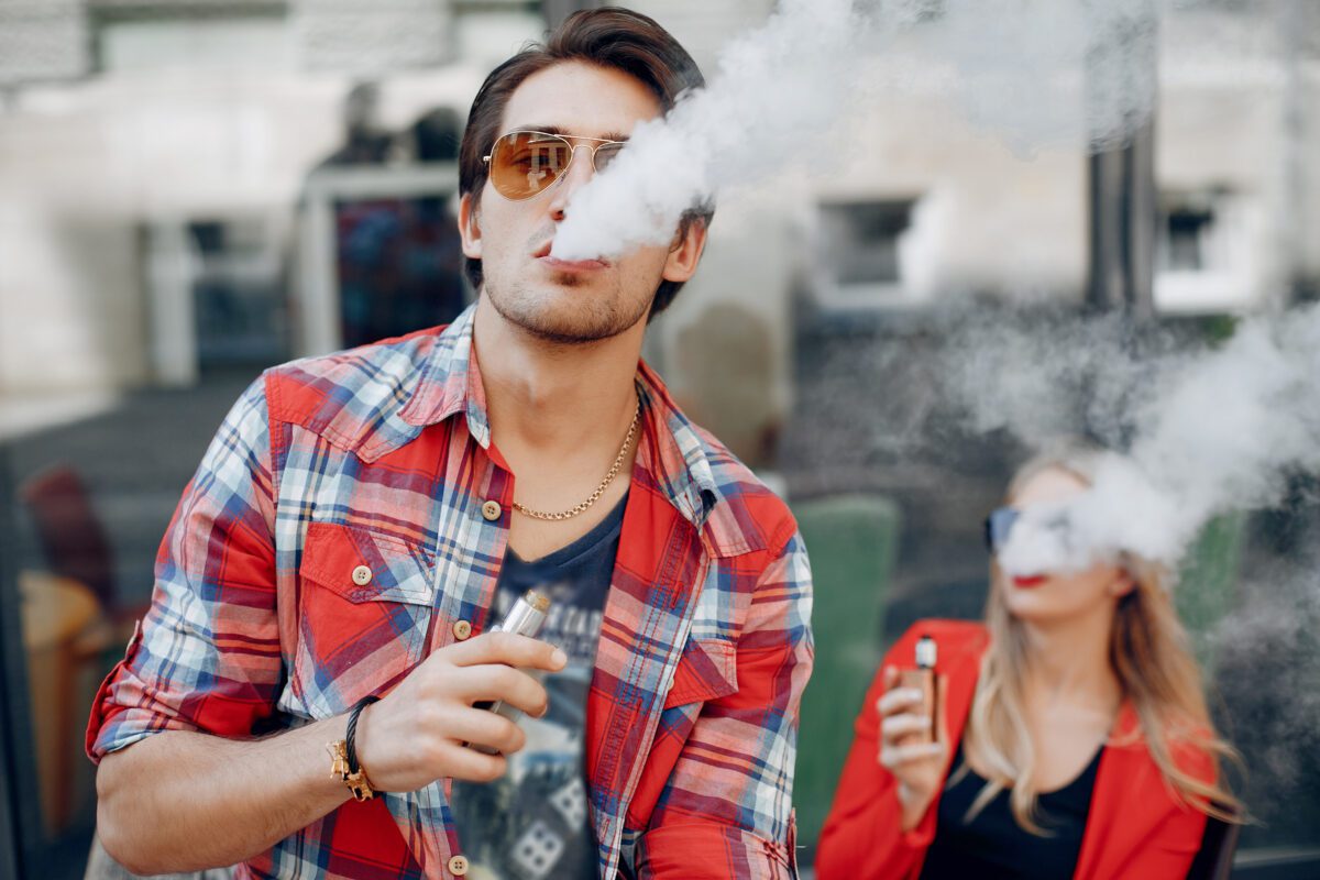 Muž a žena používajú elektronickú cigaretu na verejnom mieste, obklopení dymom.