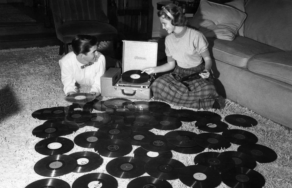 Muž a žena si púšťajú vinylové platne na prenosnom prehrávači, obklopení zbierkou LP diskov.