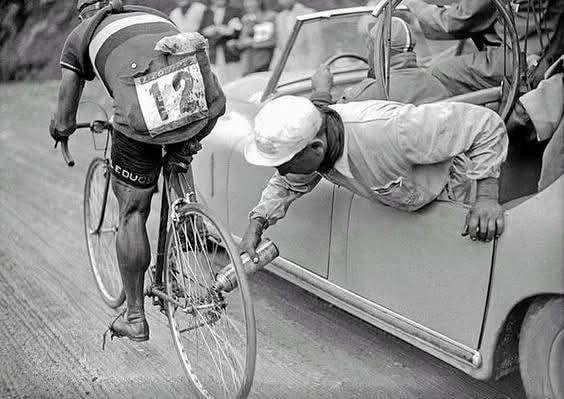 Cyklista dostáva asistenciu z auta počas pretekov. Starobylý moment zachytený na čiernobielej fotografii.