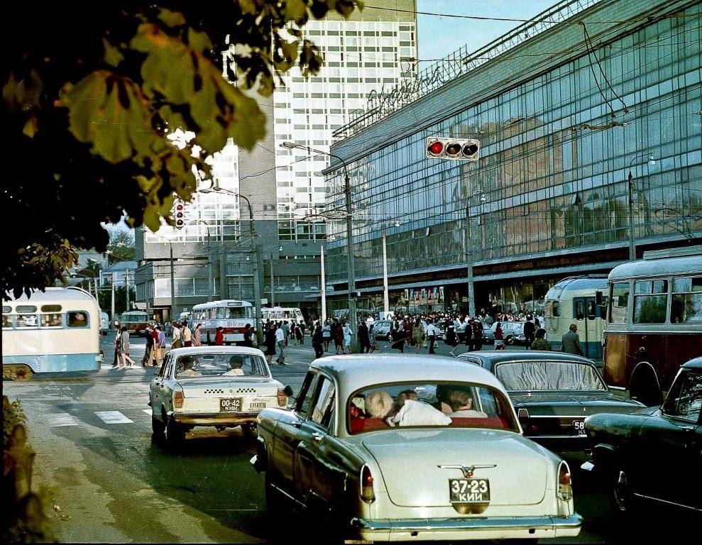 Mestská križovatka s rušnou dopravou a chodcami pred modernou presklenou budovou za slnečného dňa.
