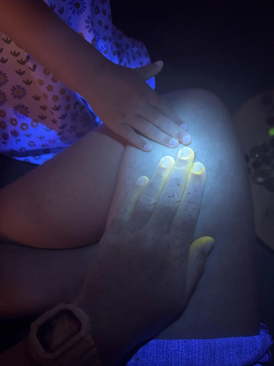 Ruky dieťaťa a dospelého svietia pod UV svetlom, dotýkajúc sa kolena v tme.