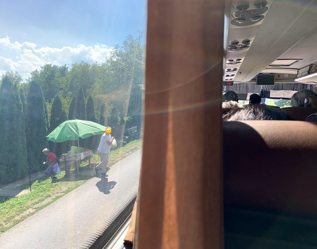 Pohľad z autobusu na ľudí predávajúcich ovocie pod zeleným dáždnikom pri ceste, slnečný letný deň.