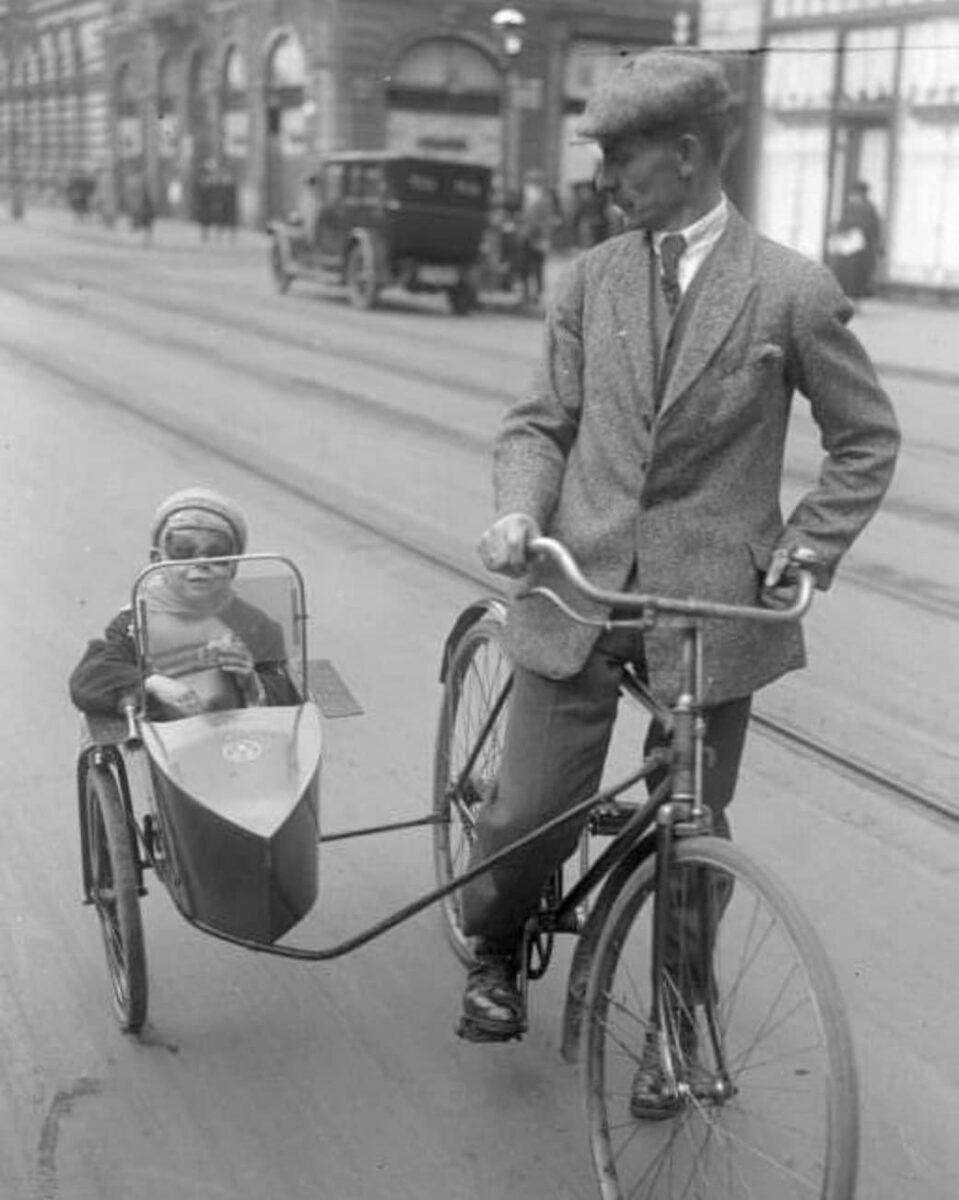 Muž bicykluje s dieťaťom v prívese po meste, čiernobiela fotografia z historického obdobia.