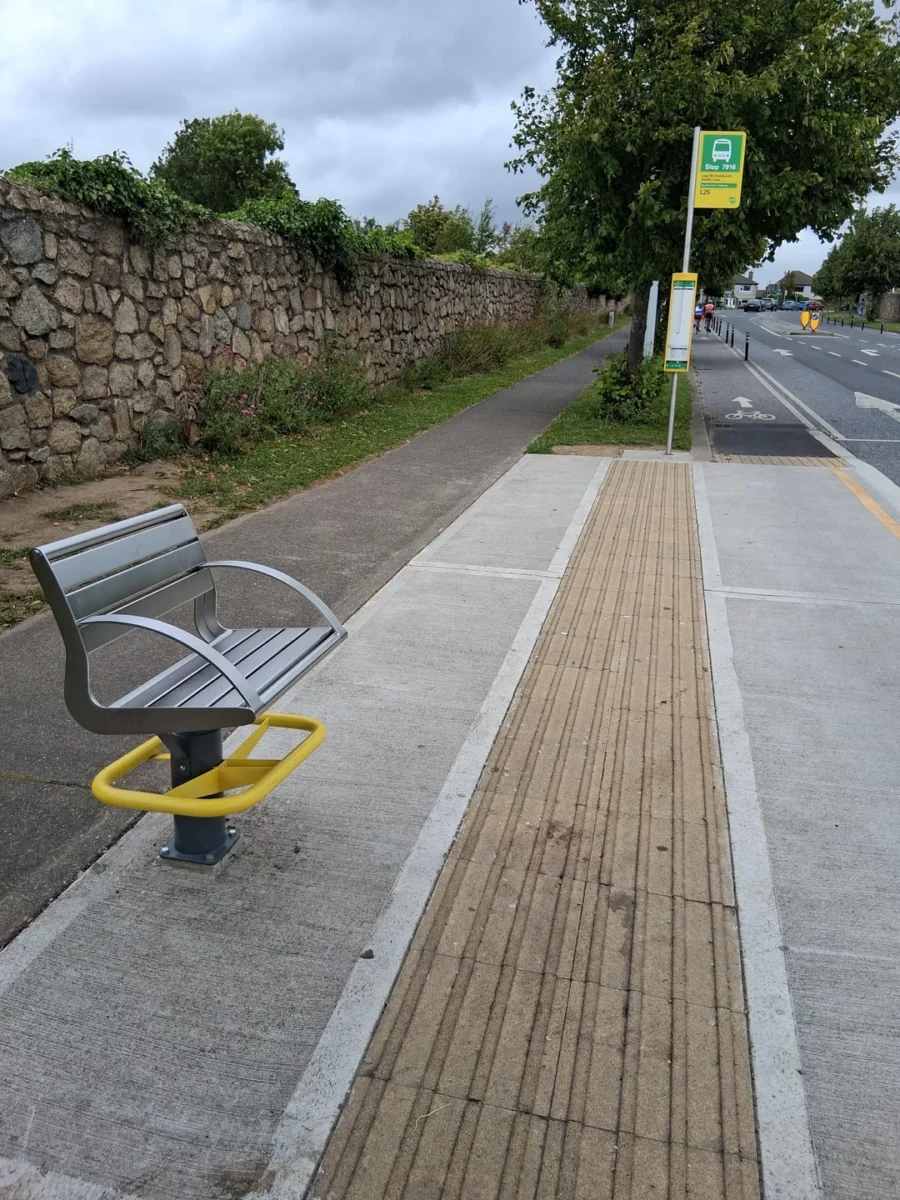 Autobusová zastávka pri kamennom múre so sedadlom bez chrbta a cyklistickým chodníkom na kraji cesty.