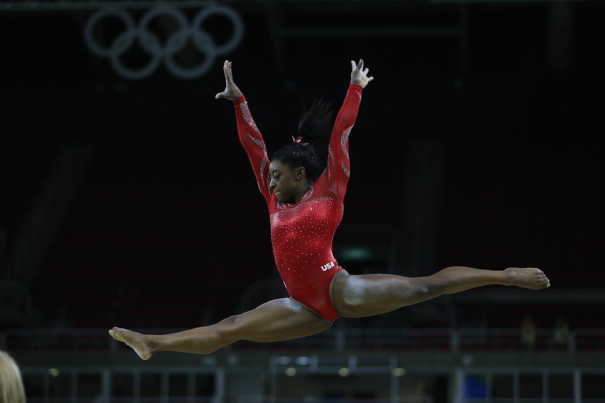 Žena vo vzduchu počas gymnasticého skoku, oblečená v červenom dresíku s olympijskými kruhmi v pozadí.