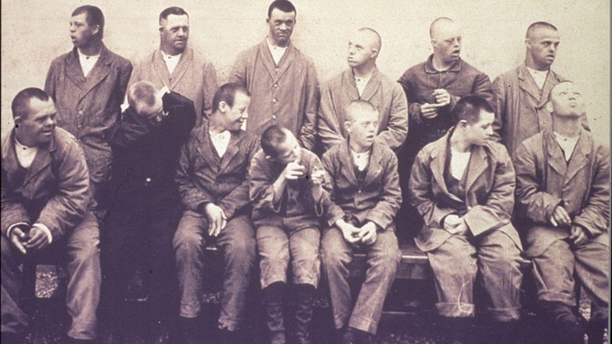 Skupinová fotografia mužov v staršom oblečení, niektorí sediaci, na historickom pozadí.