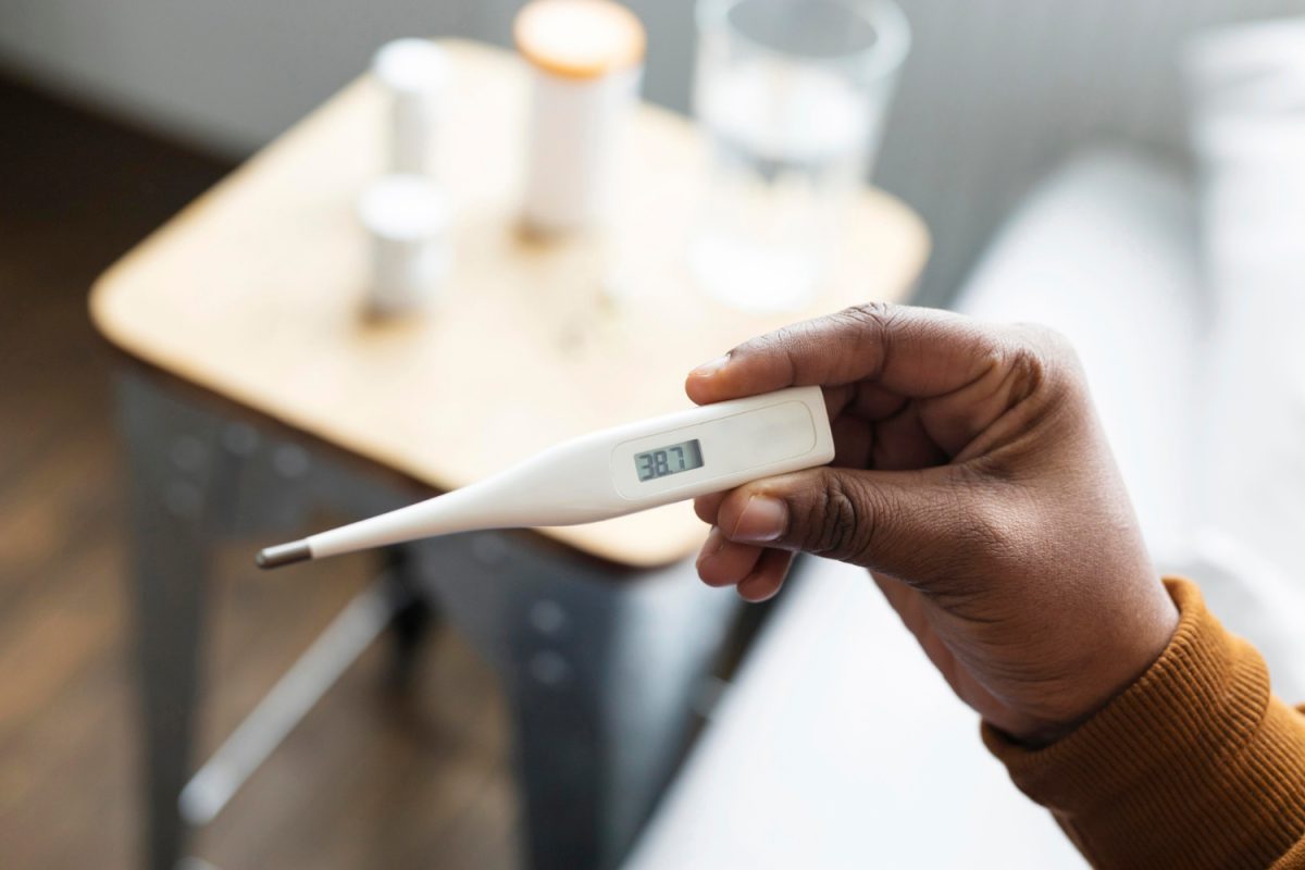 Osoba drží digitálny teplomer ukazujúci 38,7 °C na pozadí stolíka s pohárom vody.