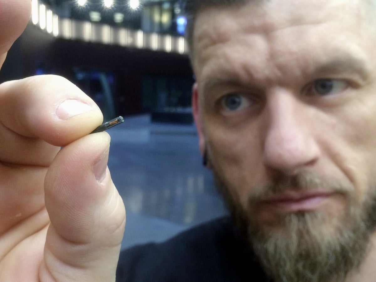 Muž drží malý implantovateľný mikročip medzi prstami, rozmazaná technologická miestnosť v pozadí.