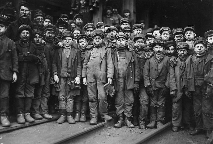 Skupina mladých chlapcov v pracovných odevoch pózujúcich pred priemyselnou budovou, historická fotografia.
