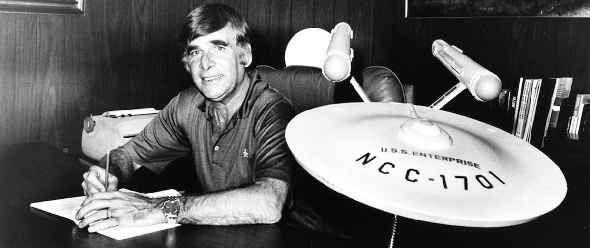 Muž pri písacom stole so starožitným modelom vesmírnej lode Enterprise NCC-1701 na pozadí.