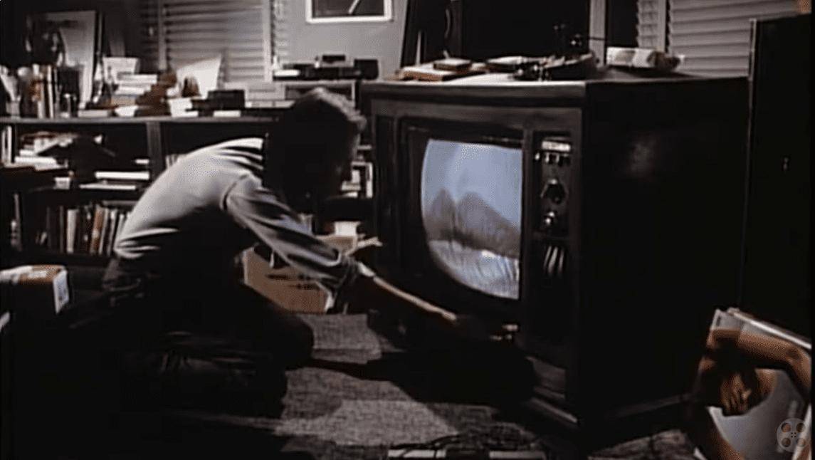 Osoba upravuje starú televíziu s obrazom úst v tmavej miestnosti plnej kníh.