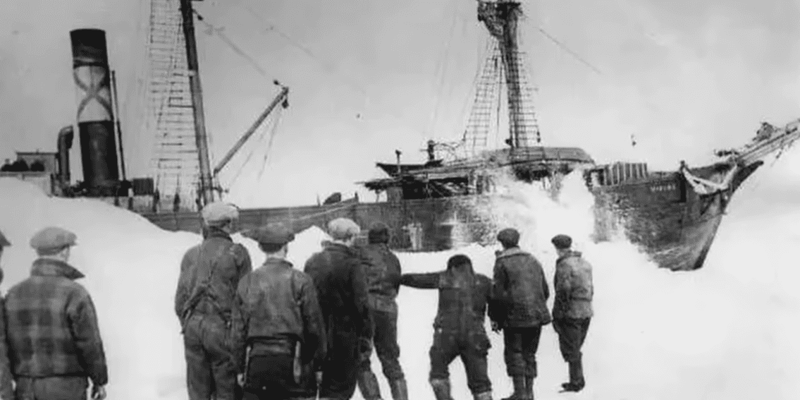 Skupina mužov pozoruje loď uviaznutú v ľade, historická čiernobiela fotografia.