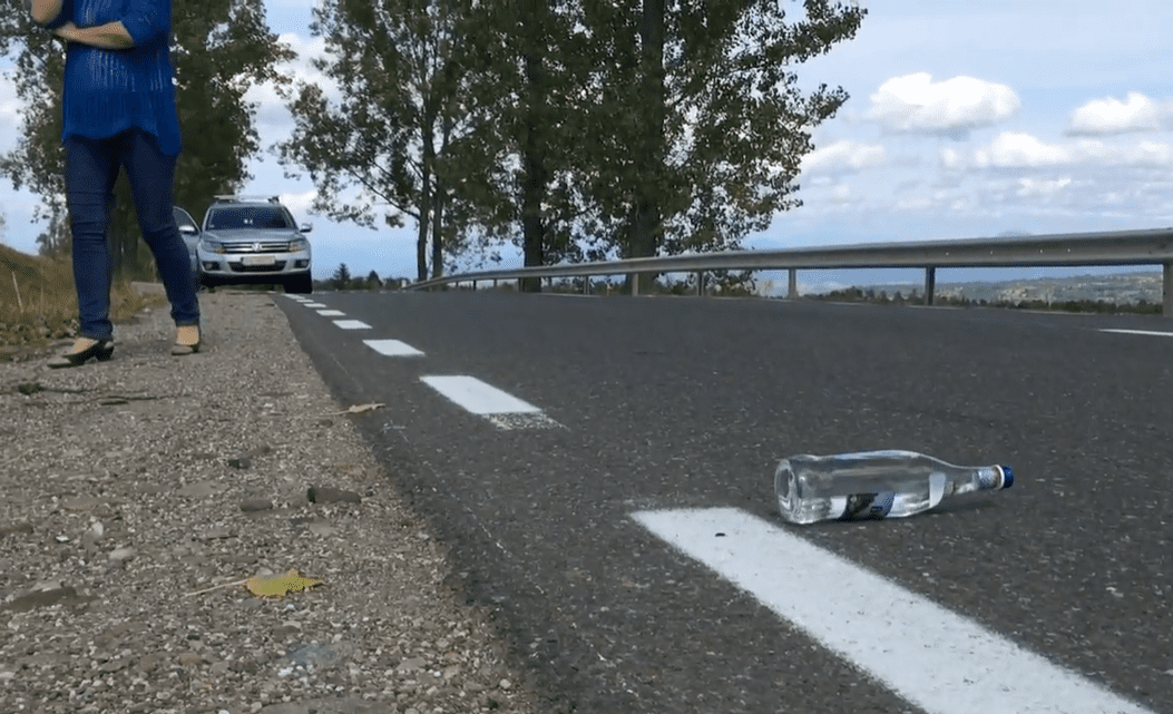 Osoba stojí vedľa cesty s fľašou na vozovke, auto na pozadí, letný deň, stromy okolo.