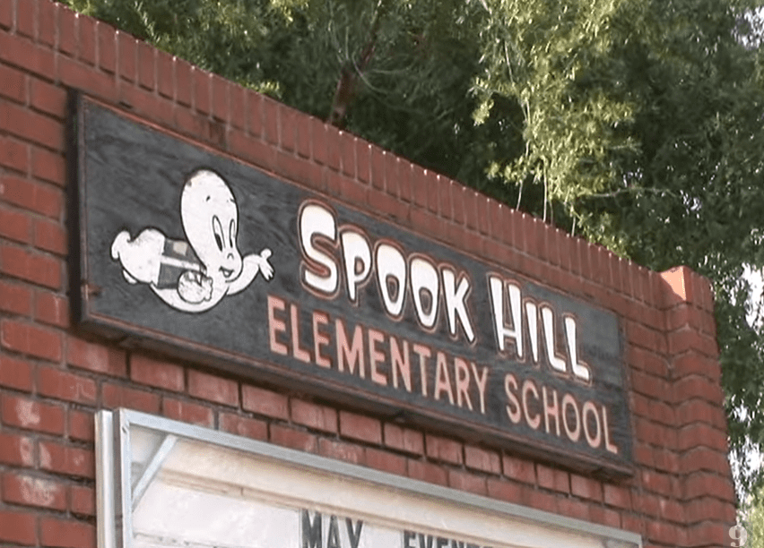 Nápis školy Spook Hill Elementary School s obrázkom kresleného ducha na tehlovej stene, stromy v pozadí.