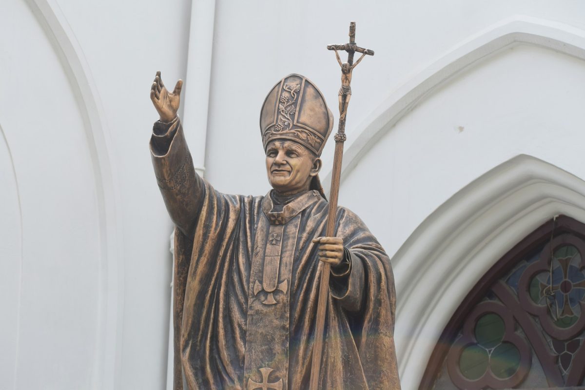 Socha náboženskej osobnosti držiaca kríž pred gotickou architektúrou.