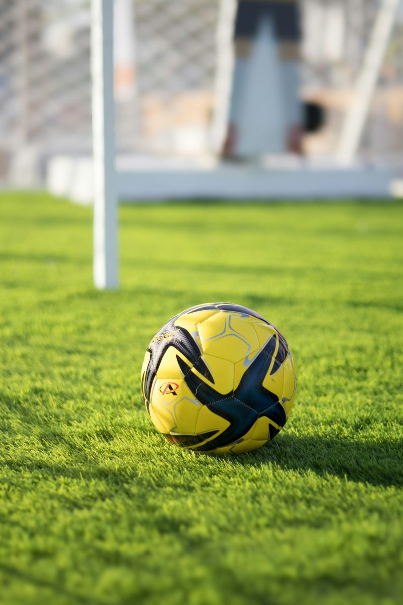 Žltá futbalová lopta na zelenej tráve pri futbalovej bránke na slnečnom ihrisku.