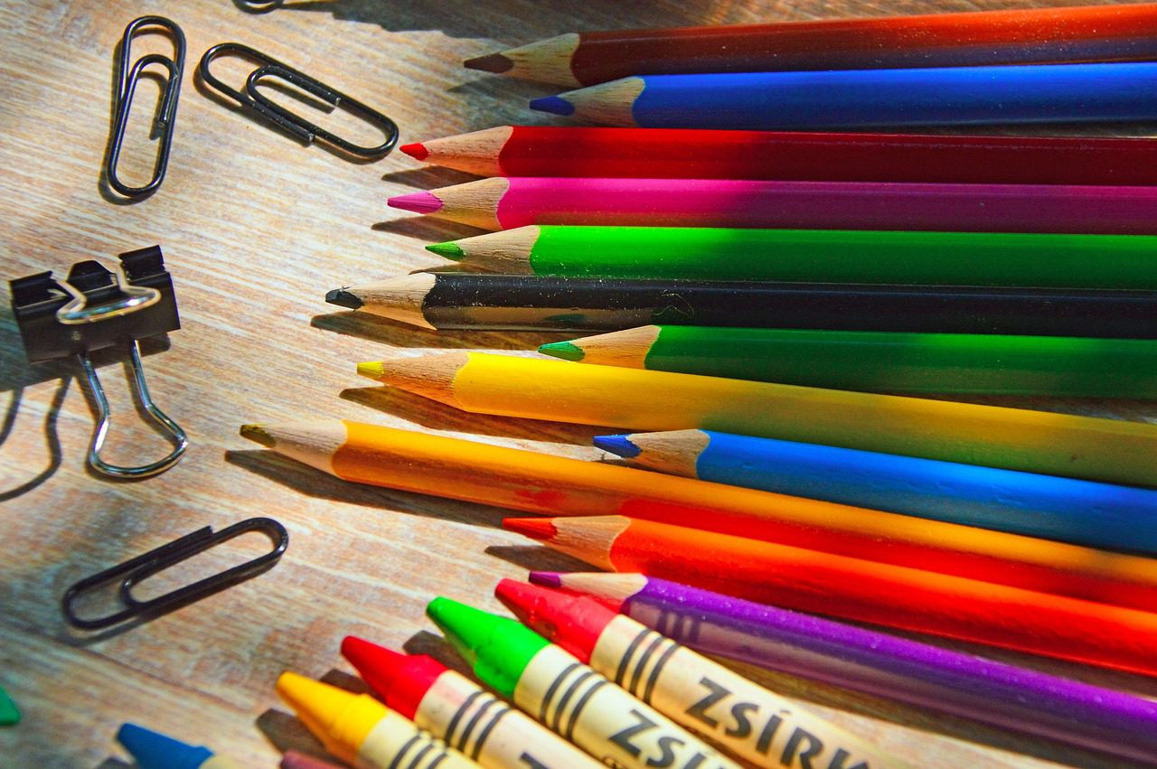 Farebné ceruzky a pastelky s kancelárskymi spinkami na stole pod slnečnými lúčmi.