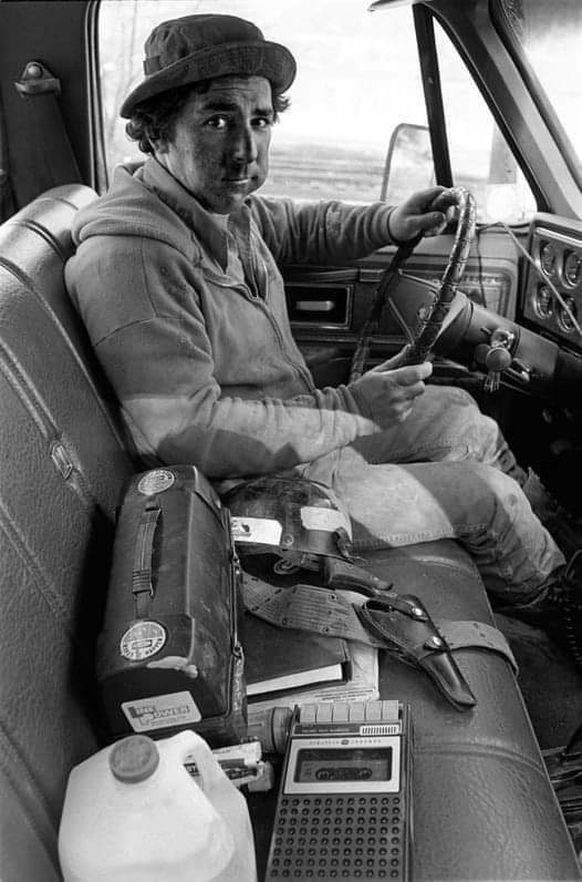 Muž sedí v aute s volantem, vedľa neho náradie a prenosný magnetofón. Vintage scéna bežného dňa.