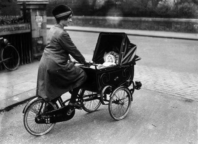 Žena na bicykli s kočíkom, v ktorom sa nachádza dieťa, na historickej ulici.
