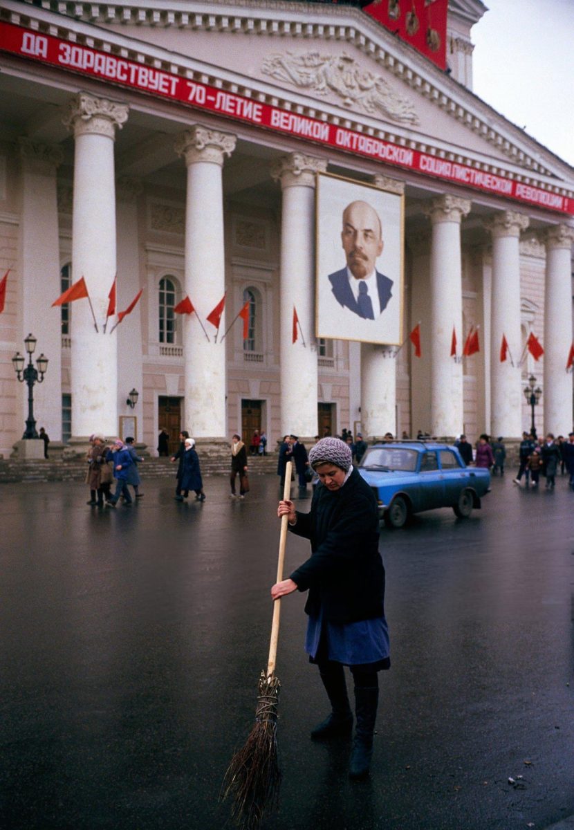 Žena zametá ulicu pred budovou s portrétom na oslavu výročia socialistickej revolúcie.