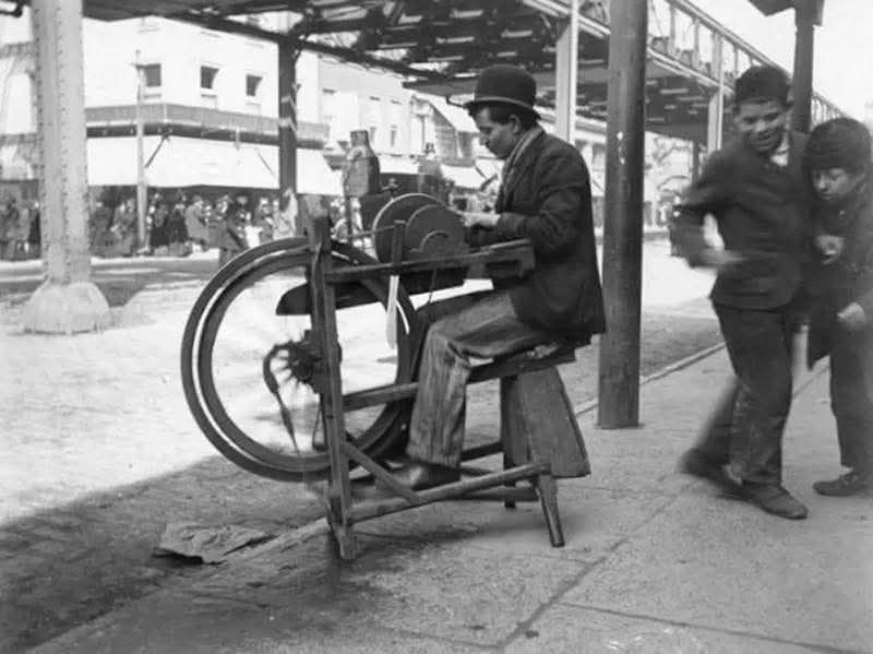 Starý remeselník ostrí nože na ručnom brúsiči na rušnej ulici v historickom meste.