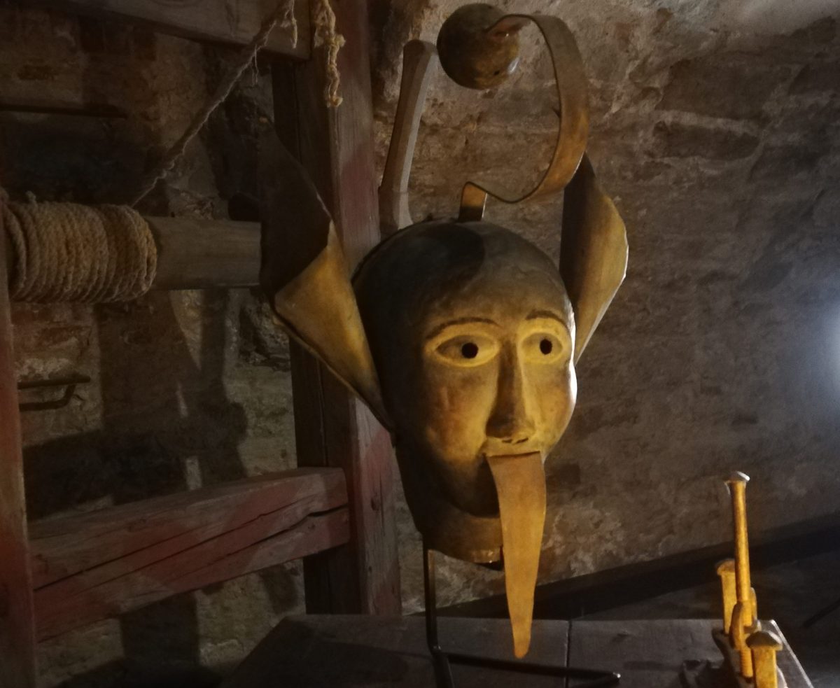 Starodávna kovová maska s dlhým nosom vystavená v kamennej sieni, historický artefakt z minulosti.