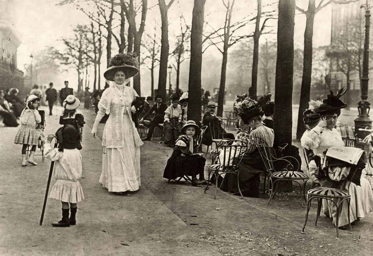 Historická promenáda s módne oblečenými ľuďmi v parku, sedí a prechádza sa medzi stromami.