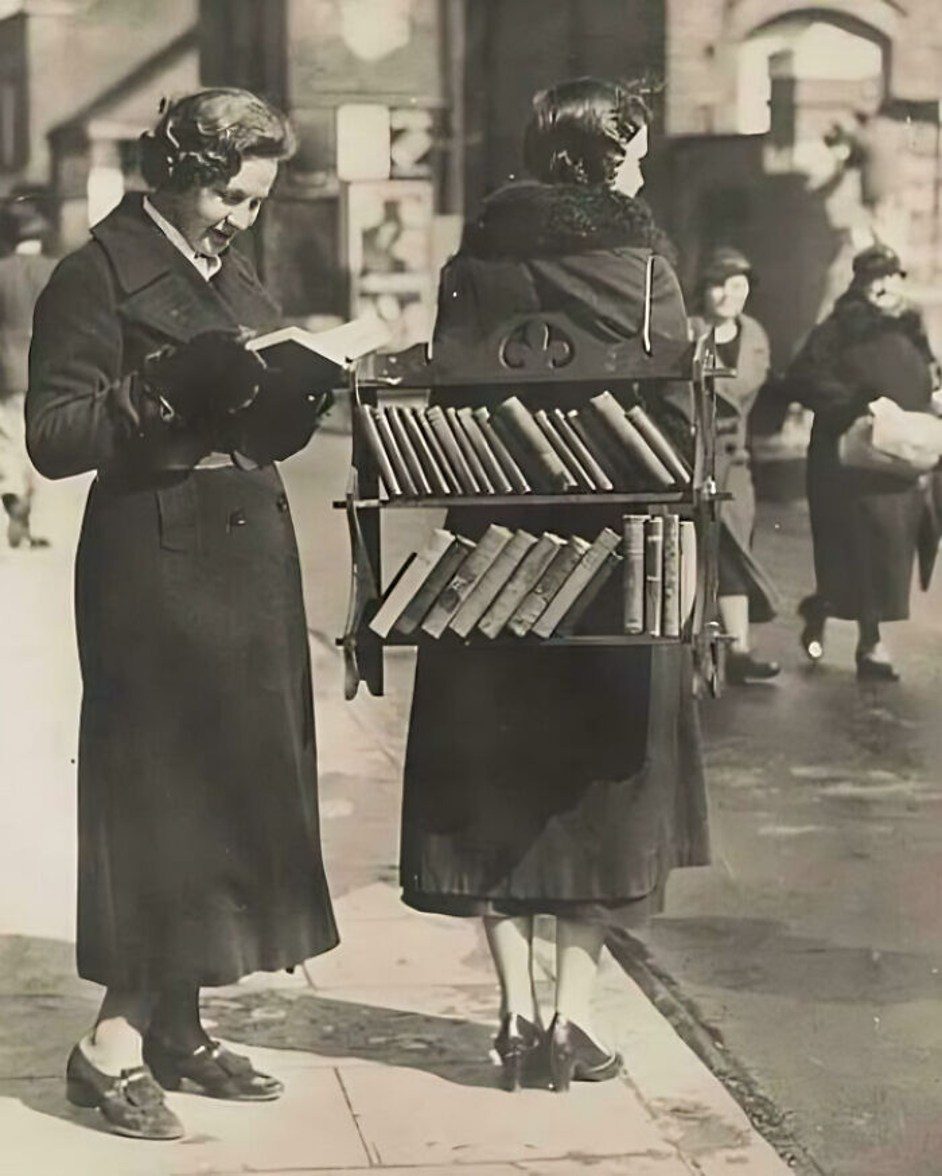 Žena stojí v ulici s prenosnou knižnicou na chrbte, druhá číta knihu. Retro fotografia.