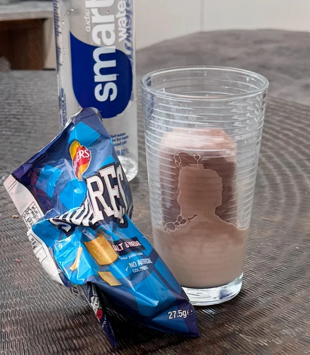 Fľaša vody, pohár s nápojom a balík čipsov na stole.