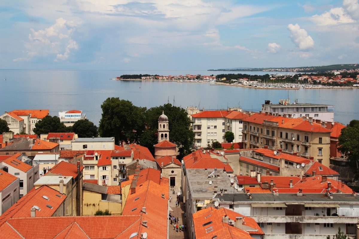 Pohľad na pobrežné mesto s červenými strechami a historickým kostolom, v pozadí modré more a zeleň.