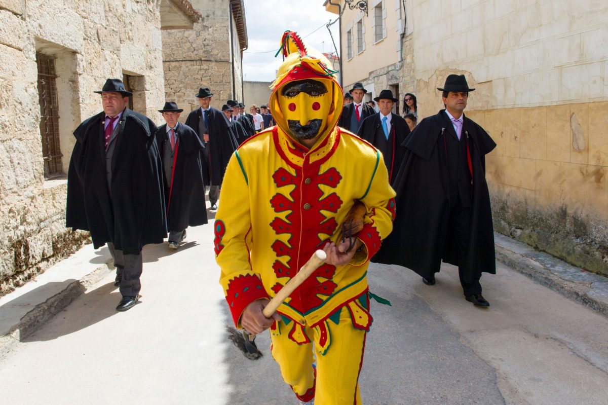 Muž v žltom kostýme a maske vedie sprievod ľudí v klobúkoch v historickej mestskej uličke.