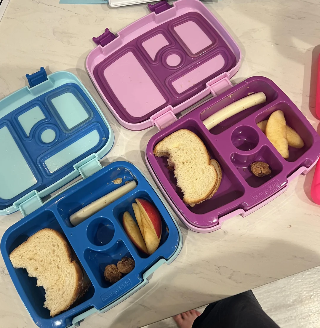 Tri otvárateľné lunchboxy s rôznymi snackmi: sendviče, jablká, syr a malé pochutiny na mramorovom povrchu.