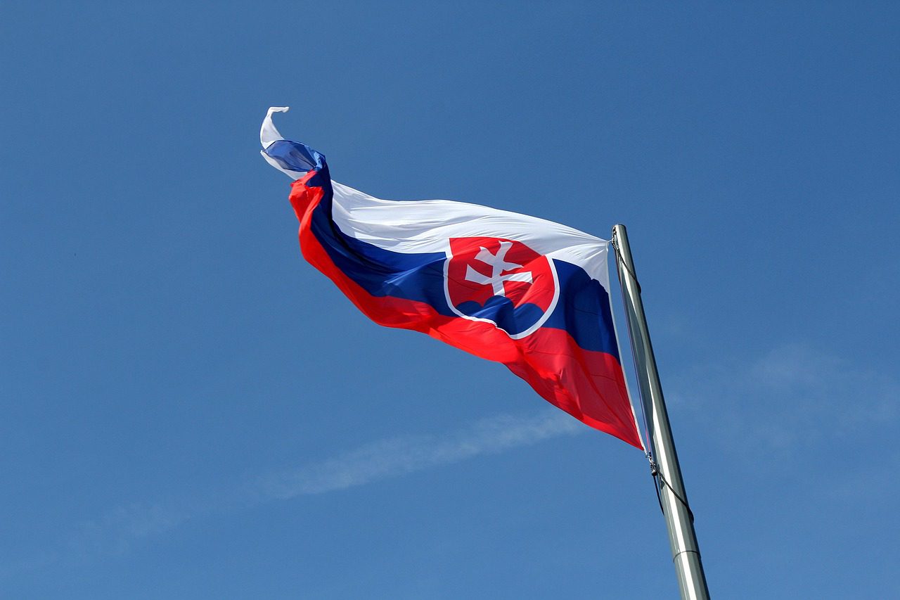 Slovenská vlajka vejúca na stožiari proti modrej oblohe.