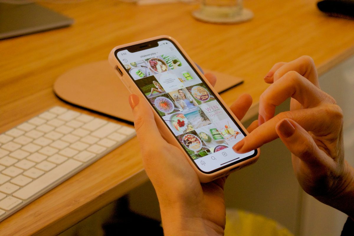 Osoba prehliada Instagram v mobile na drevenom pracovnom stole s klávesnicou a notebookom.