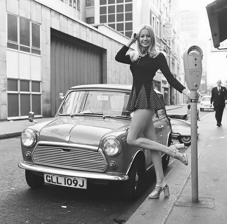 Žena opretá o parkovací automat vedľa historického Mini Cooper vo vintage mestskej ulici.