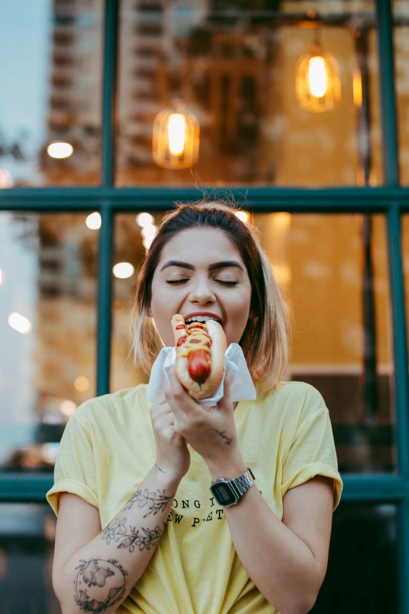 Žena si pochutnáva na hotdogu pred oknom s jasnými svetlami v pozadí.