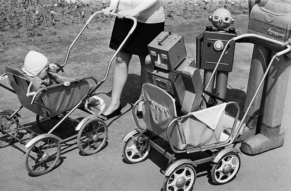 Žena tlačí kočík s bábätkom a vedľa je kočík s robotom na ulici, čiernobiela fotka.
