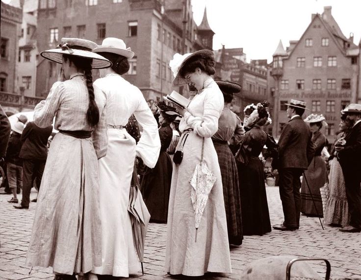 Historická ulica plná ľudí v dobových kostýmoch, ženy čítajúce knihu a držiace dáždniky.
