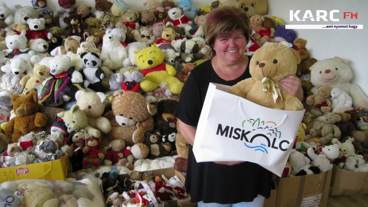 Žena drží plyšového medvedíka a tašku pred zbierkou hračiek, usmievajúc sa.
