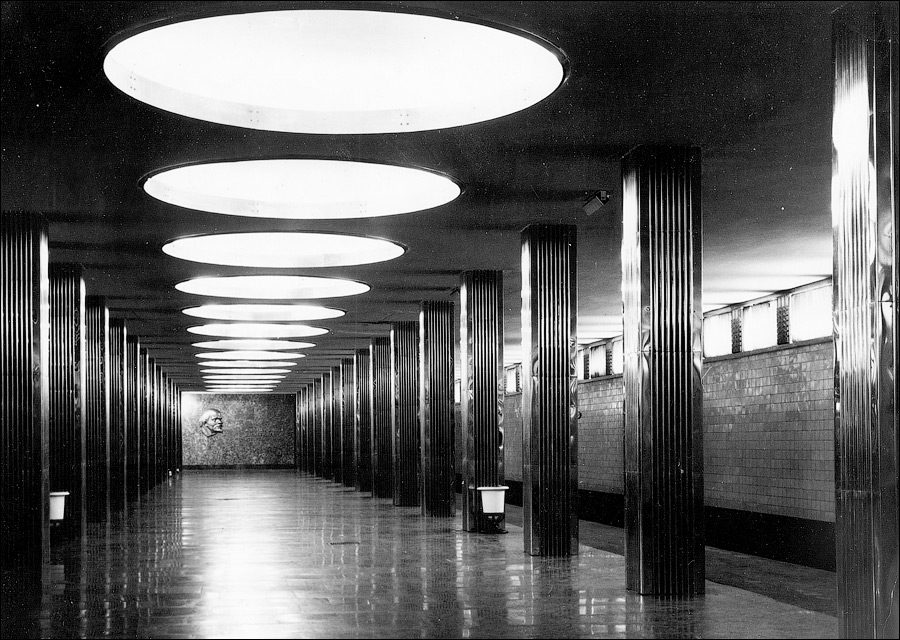 Čiernobiela fotografia chodby s modernými stĺpmi a okrúhlym osvetlením na strope.