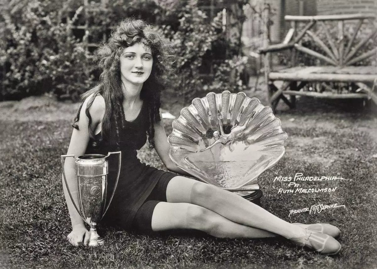 Žena sedí na trávniku vedľa dvoch veľkých trofejí, oblečená v dobovej móde.