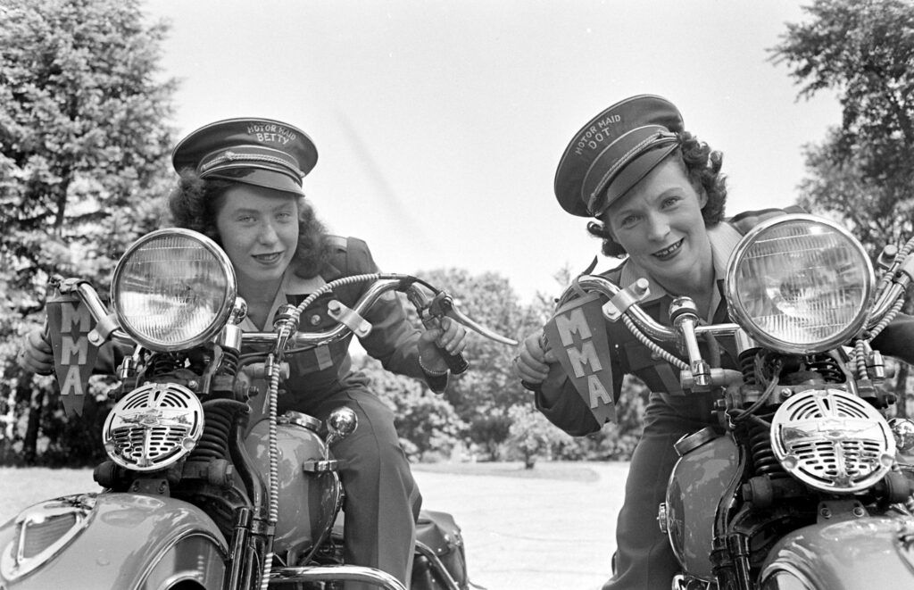 Dve ženy na cestných motocykloch v uniformách s úsmevom pozerajú do kamery, pripravené na jazdu.