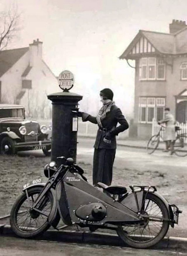 Žena v kabáte posiela list do poštovej schránky, vintage motorka a auto v pozadí. Vintage ulicné prostredie.