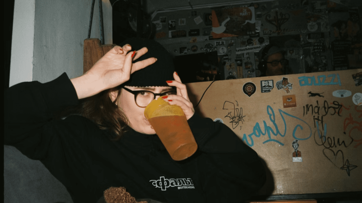 Osoba v čiernej čiapke pije nápoj v pohári, bar s nálepkami na pozadí.