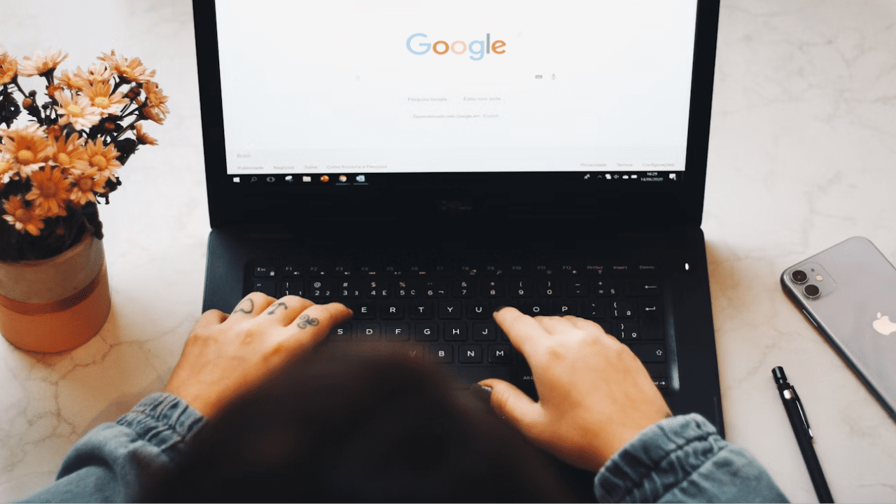 Osoba píše na notebooku s otvorenou stránkou Google, vedľa je položený telefón a váza s kvetmi.