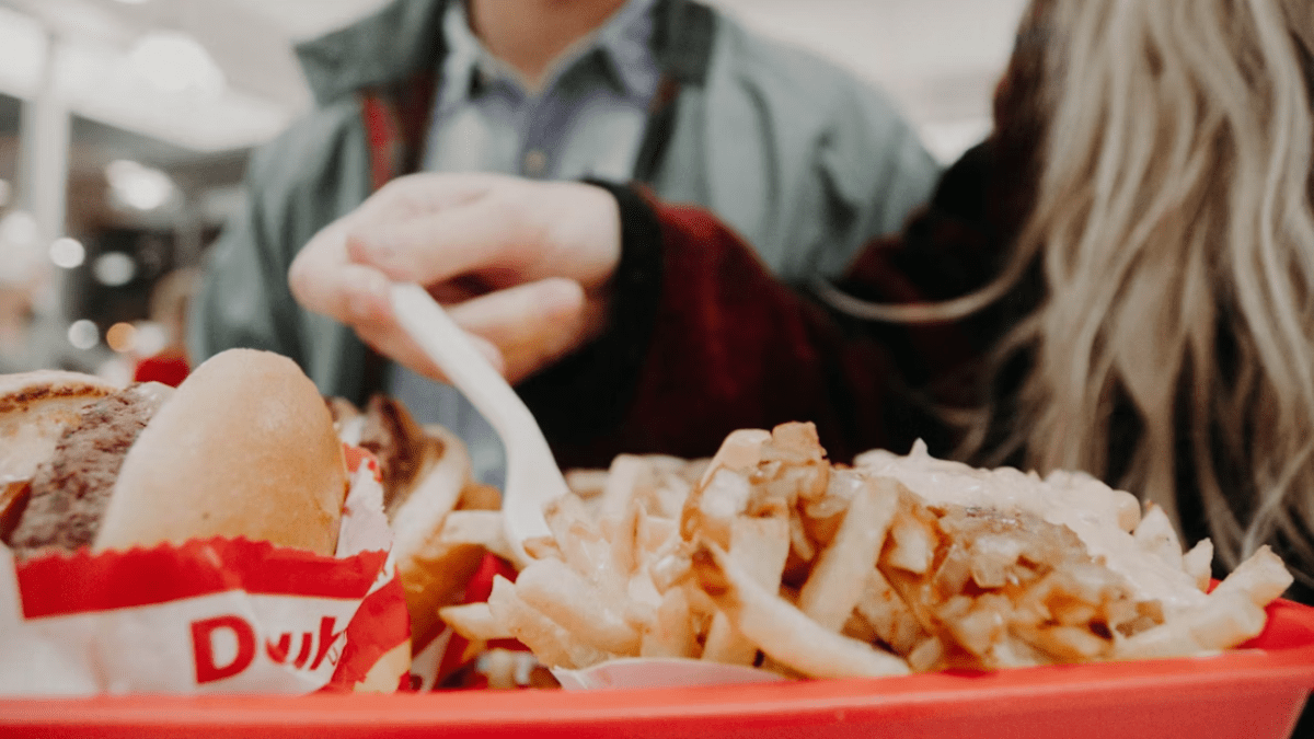 Osoba konzumujúca hamburger a hranolky z červeného podnosu vo fastfoode.