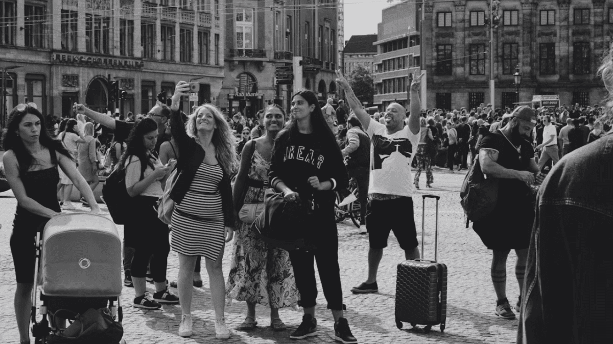 Skupina turistov na rušnom námestí robí selfie v slnečný deň, okoloidúci s kočíkom a kufrom.