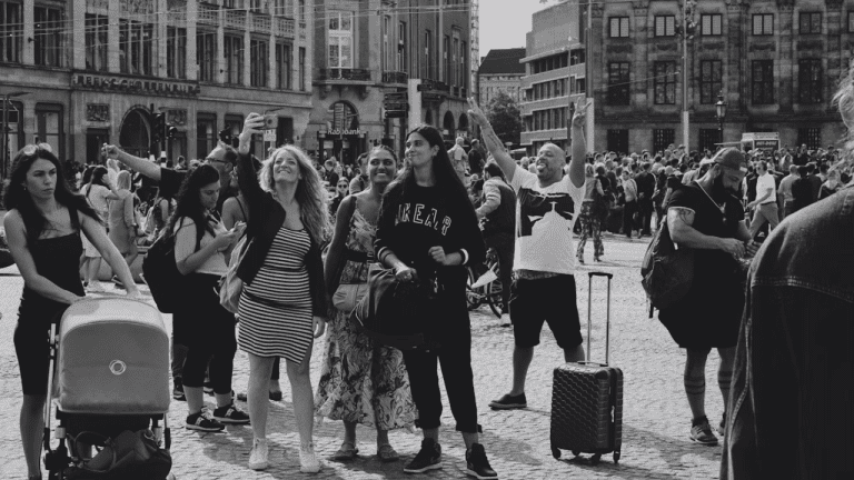 Skupina turistov na rušnom námestí robí selfie v slnečný deň, okoloidúci s kočíkom a kufrom.