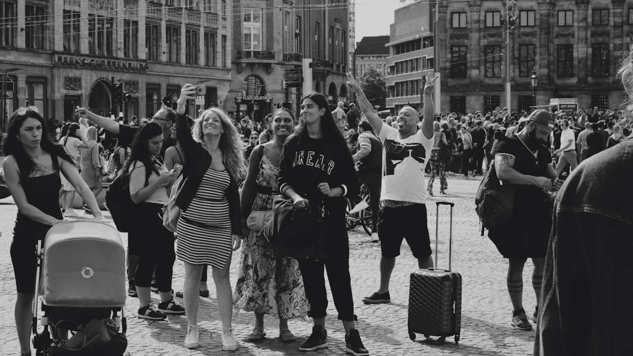 Skupina turistov na rušnom námestí robí selfie v slnečný deň, okoloidúci s kočíkom a kufrom.
