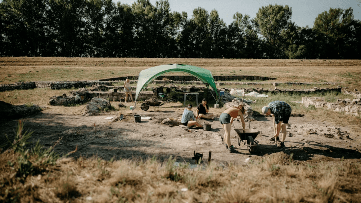 Archeologický výskum na slnečnom mieste, ľudia pracujú pod zeleným stanom, nástroje a vozík v teréne.